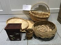 Lot photograph showing the wooden mortar and pestle, rectangular wooden container with mother of pearl bird inlays, woven basket with handle, round basket tray, flat bamboo tray, and round bamboo basket grouped together.
