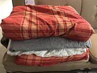 Stack of king size bedding including comforter and multiple comforter covers folded on a couch, showing red and beige plaid patterned comforter on top.
