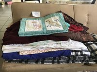 Photo of a stack of various bedding items including comforter covers, linen, blankets, and quilts arranged on a couch, showing material color and texture variety