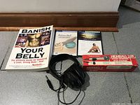 Photo showing five items: the 'Banish Your Belly' book, 'Balance Ball' exercise DVD, 'Yoga in Practice' exercise DVD, a box of Highball 2 tennis balls, and wired headphones.
