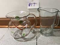 Two glass pitchers side by side; one round with orange and green swirled glass decoration, one taller plain clear glass pitcher.