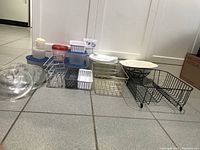 Photo of various plastic labeled food storage containers with blue and red lids, metal and plastic kitchen racks, glass lids, white ceramic bowl, and black metal stands all arranged on tiled floor.