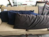 Three long and body pillows on beige couch, two with navy blue pillowcases and one with brown pillowcase.