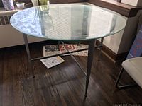 Round glass table with chrome base shown on wooden floor near window.