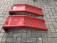 Pair of red metal car ramps placed side by side on outdoor paving, showing their shape and perforations.