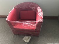 Red tub-style chair wrapped in protective plastic with matching back pillow visible inside the seat area. Chair is on carpeted floor near wall.