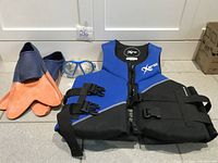 Photo showing the blue and black life jacket vest, a pair of orange and blue fins, and swimming goggles arranged on floor.