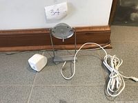Photo showing the small LED desk lamp with circular light head, silver finish, and white rectangular base, with power cord and adapter on the floor.