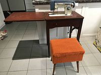 Full view of the vintage sewing machine mounted on a dark wooden sewing table with an extension leaf, alongside an orange upholstered storage stool.
