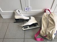 Full view of the pair of white women's figure skates size 9 next to the white carrying bag with pink straps.