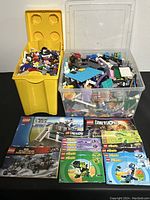 View of two storage containers, one yellow LEGO bin and one clear plastic box with assorted LEGO pieces and instruction manuals laid out in front