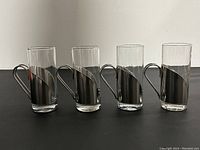 Front view of four stainless steel and glass espresso mugs arranged in a row with handles to the side.