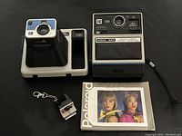 Photo showing Kodak The Handle camera, Kodak EK4 instant camera, manual, and keychain in one frame on black surface.