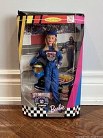Front view of the unopened boxed 1998 50th Anniversary NASCAR Barbie Doll showing the doll in blue racing jumpsuit holding a helmet with background racing scene and box branding.