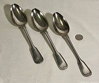 Six large antique sterling silver soup spoons arranged next to a nickel coin for size reference on a light fabric surface. Spoons show patina and tarnish.