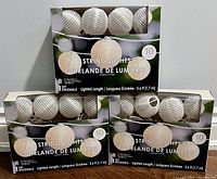 Three boxes of Ashland fabric lantern string lights in original packaging, showing 10 white fabric lantern lights per box on 5.6 ft cord.