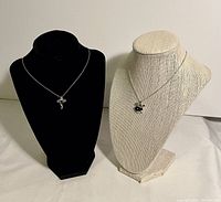 Two necklaces displayed on black and beige bust stands side by side, showing the cross pendant on the black bust and the whale pendant on the beige bust.