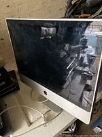 Front view of Apple iMac showing black screen with silver aluminum body and Apple logo. Screen is off, reflective and appears to have some smudges or dust.