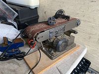 Side view of electric belt sander on wooden base with worn abrasive belt and attached power cord with non-standard plug