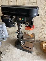 Full side view of a black bench drill showing the motor housing, column, base, and drilled table with rust spots, power switch visible on the head unit.