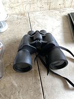 Pair of black Bushmaster binoculars showing the front lenses, central focus knob, and attached neck strap on a table.