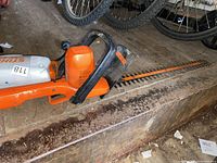 Photo of an orange and black Black and Decker electric hedge trimmer placed on a concrete surface beside bicycles.