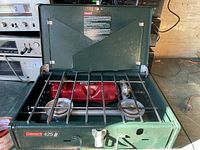 Front view of Coleman 425 two-burner camping stove with lid opened, showing two burners, red fuel tank and metal grate.