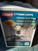 Photo of the original cardboard box featuring the Coleman propane lantern, showing product image, branding, and electronic ignition feature.