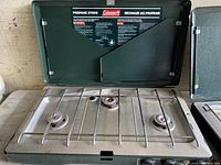 Front view of the Coleman propane stove open with three burners visible. Foldable wind guards are up, and control knobs at bottom.