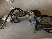 Photo of a gray and black Dremel vibrating tool showing the Dremel label and attached power cord with plug, placed on a concrete floor.