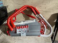Photo of folded fire escape ladder with red hooks and coiled red rope, highlighting aluminum ladder steps and attached instruction label.