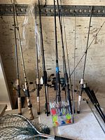 Seven fishing rods with reels leaning against wall, net and lure box at base