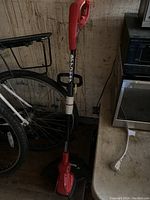 Full view of the red and black Homelite electric grass trimmer standing upright next to a bicycle and a toolbox.