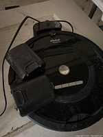 Photo showing black iRobot Roomba vacuum, charger base, and two extra battery packs placed on a surface with some dust.