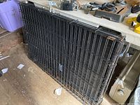 Folded large black metal wire dog crate leaning against a workbench in a workshop setting.