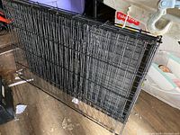 Photo of folded medium-sized metal dog crate, black wire mesh, placed on a wooden floor with miscellaneous items around.
