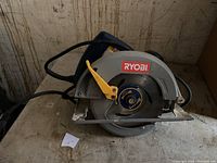 Front side view of Ryobi circular saw showing the blade, yellow adjustment lever, power cord, and Ryobi logo on the guard.