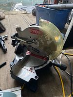 Side view of a Ryobi radial arm saw with circular blade and yellow plastic safety guard, placed on a wooden surface.