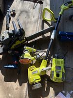 Power tools spread out on wooden surface including sander, drill, chainsaw, string trimmer, battery charger, and battery pack, all Ryobi brand, mostly yellow with black accents.