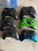 Six Xbox wireless controllers laid out on a surface: five black, one green Minecraft-themed, one metallic blue. Controllers show signs of use and are untested.