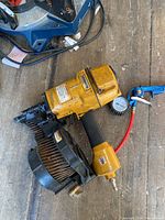 Photo shows a yellow and black Stanley-Bostich coil nailer with coil magazine, pneumatic hose connected to a pressure gauge.