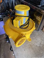 Photo showing top and side view of yellow air mover with motor and power cord wrapped around it, placed on a table with other items in background.