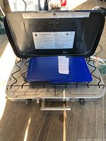 Front-top view of the Woods portable propane stove showing two burners under the open hinged lid with a blue drip tray and control knobs at the front.