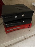 Three Xbox 360 consoles stacked; two black, one red, visible surface wear on top black console