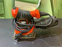 Front and top side view of Black & Decker palm sander showing ergonomic handle, ventilation slots, dust bag, and wrapped power cord.