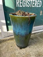 Full view of ceramic jardiniere showing green to turquoise gradient glaze with dried plants inside, placed on concrete ground with signage background.