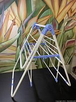 Full view of the vintage folding laundry drying rack showing the A-frame design and multiple rods for hanging clothes, against a backdrop with large green and orange plant imagery.