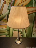 Burnished metal table lamp on black surface with beige pleated fabric shade, set against a painted background.
