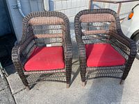 Pair of brown faux wicker patio chairs with curved back and armrests. Both chairs have removable red cushions on the seats. Shown outdoors on concrete.