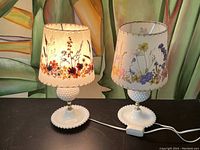 Pair of vintage hobnail milk glass table lamps with floral pressed flower fabric shades shown side-by-side on black surface with painted plant background.
